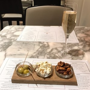 A glass of champagne and a wooden tray with three small dishes of snacks: green olives, savoury popcorn and spiced nuts. Set on a marble-topped dining table.
