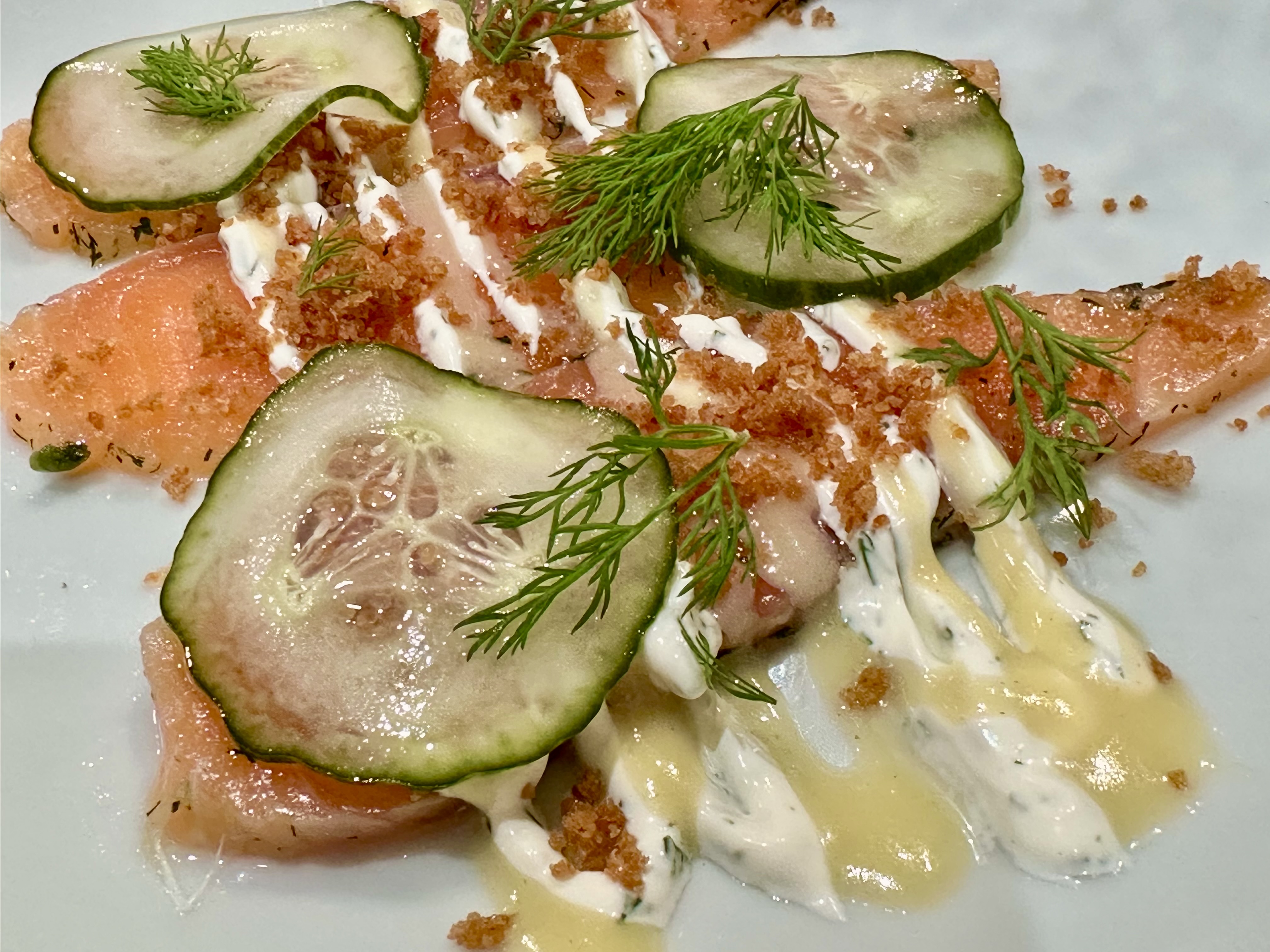 Slices of chilled, cured salmon arranged on a plate with a garnish of thinly sliced pickled cucumber, two different dressings, sprigs of dill and golden crumbs.