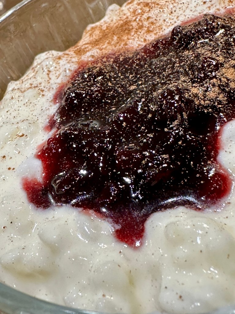 A close-up image of glistening, thick rice pudding with a pool of dark red berry jam. A sprinkling of ground cinnamon can be seen garnishing the dish.